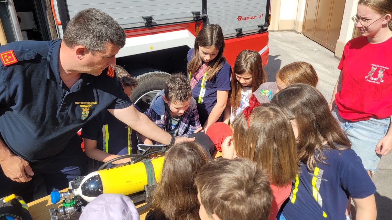 Ein Feuerwehrmann zeigt einem Jungen ein Gerät, während Kinder aufmerksam davor stehen und ein Feuerwehrauto im Hintergrund ist.