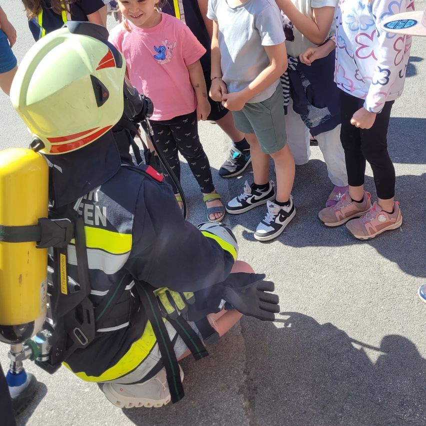 Ein Feuerwehrmann in voller Ausrüstung kniet auf dem Boden und unterhält sich mit Kindern in Freizeitkleidung an einem sonnigen Tag.