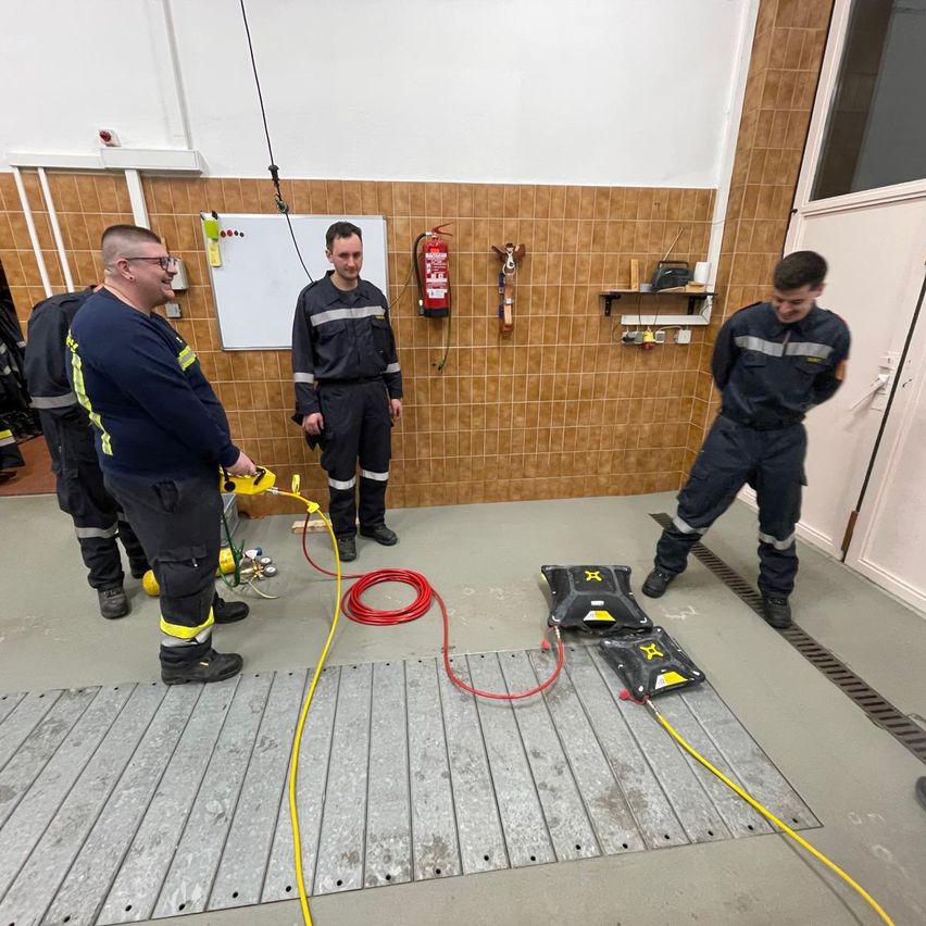 Eine Gruppe von Feuerwehrleuten befindet sich in einem Schulungsraum. Einer hält ein gelbes Werkzeug. Ein roter Schlauch liegt auf dem Boden. Ein anderer lehnt sich an eine Tür. Eine Tafel und ein Feuerlöscher sind an der Wand angebracht.