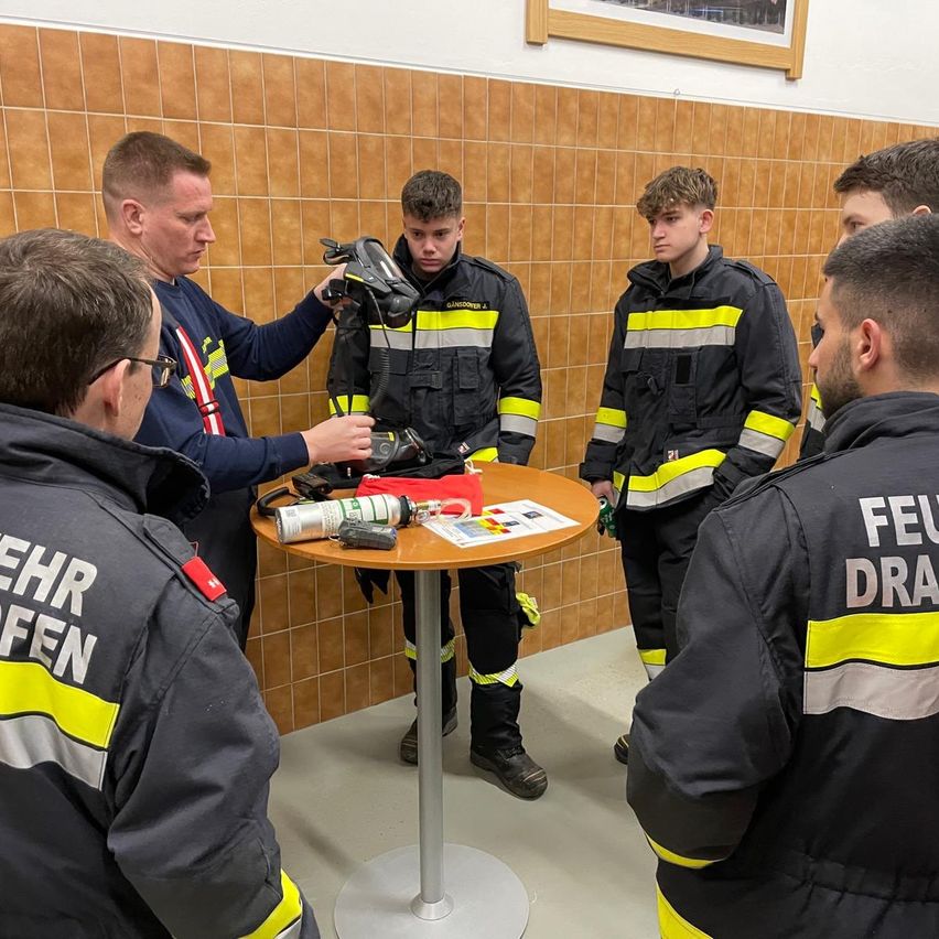 Eine Gruppe von Feuerwehrleuten versammelt sich um einen Tisch. Ein Mann hält einen Helm, und verschiedene Ausrüstungsgegenstände liegen auf dem Tisch.