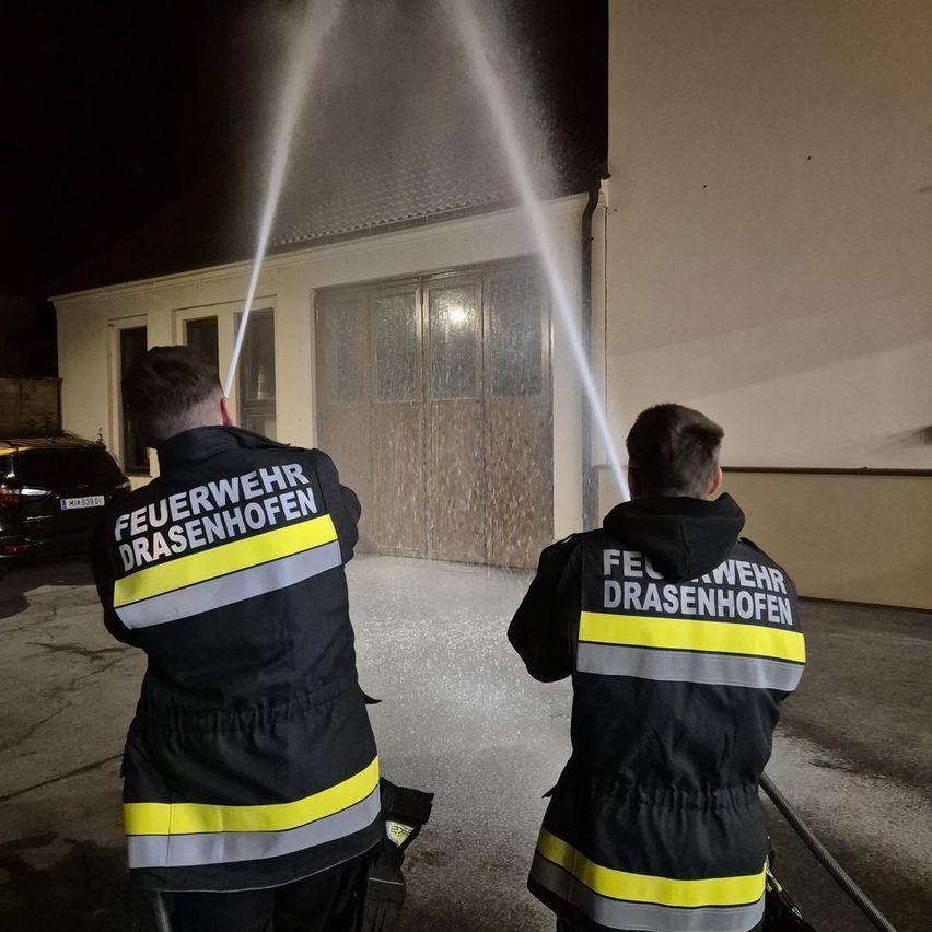 Zwei Feuerwehrleute in Uniform sprühen nachts Wasser auf ein Gebäude.