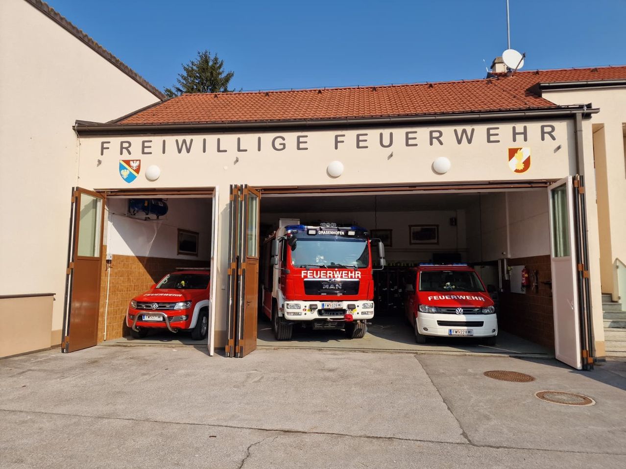 Das Freiwillige Feuerwehr Gebäude hat seine Türen geöffnet und zeigt zwei rote Feuerwehrfahrzeuge, die drinnen geparkt sind. An der Wand befindet sich ein Logo und der Schriftzug 'Freiwillige Feuerwehr'.