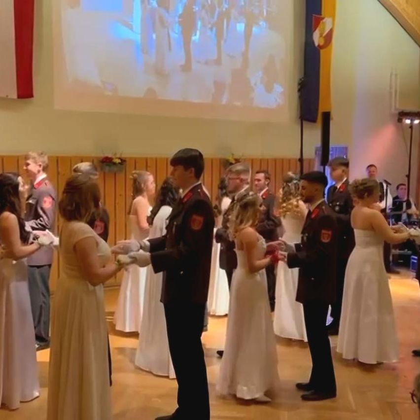 Eine Gruppe von Menschen in Militäruniformen versammelt sich in einem Saal. Zwei Paare werden in einer Zeremonie geehrt. Sie tauschen Ringe aus.