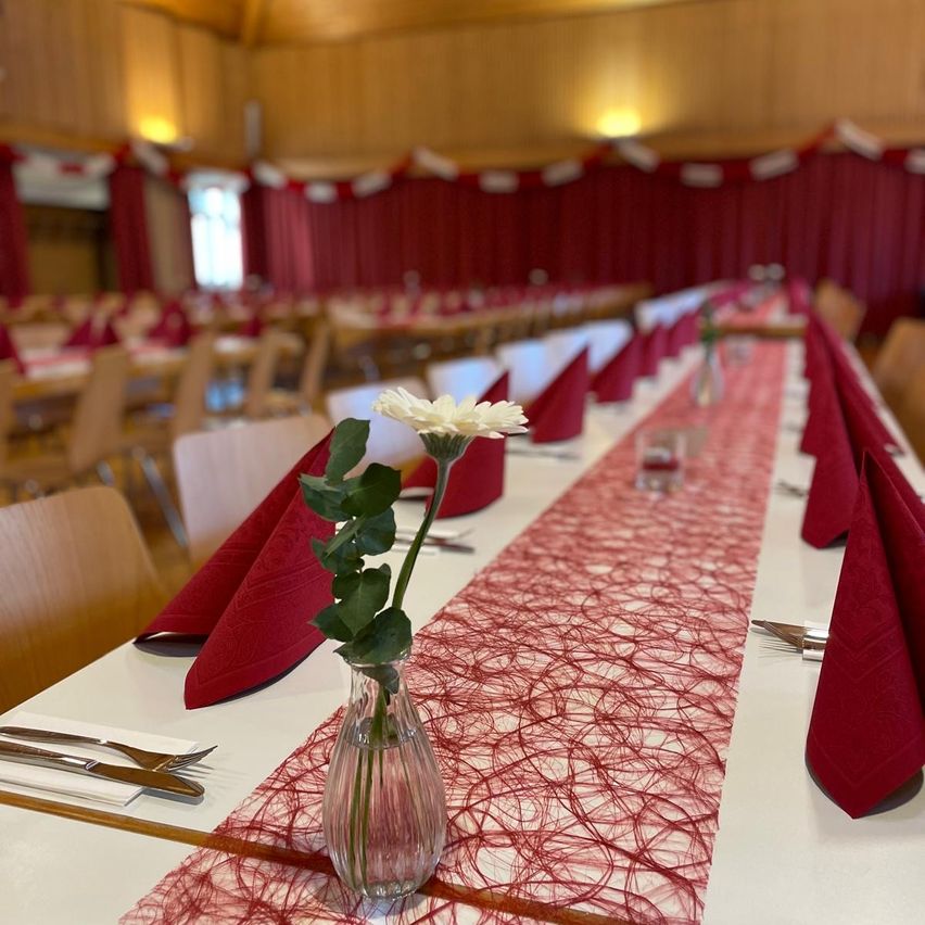 Ein Speisesaal ist für eine formelle Veranstaltung vorbereitet. Lange Tische sind mit roten Tischdecken und Servietten geschmückt. Ein Mittelstück mit einer weißen Blume in einer klaren Vase verleiht dem Raum einen Hauch von Eleganz.