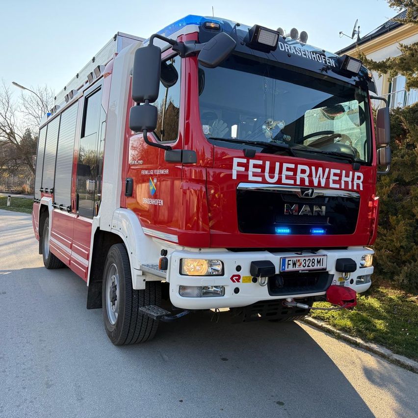 Ein roter Feuerwehrwagen mit der Aufschrift 'FEUERWEHR' steht am Straßenrand. Er hat ein blaues Licht oben und ein Kennzeichen 'FW 328 MI'.