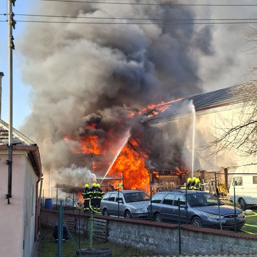 Feuerwehrleute bekämpfen ein großes Feuer, das ein Gebäude umgibt. Rauch und Flammen erheben sich über die Struktur. Zwei Autos sind in der Nähe geparkt, und weitere Fahrzeuge sind im Hintergrund sichtbar.