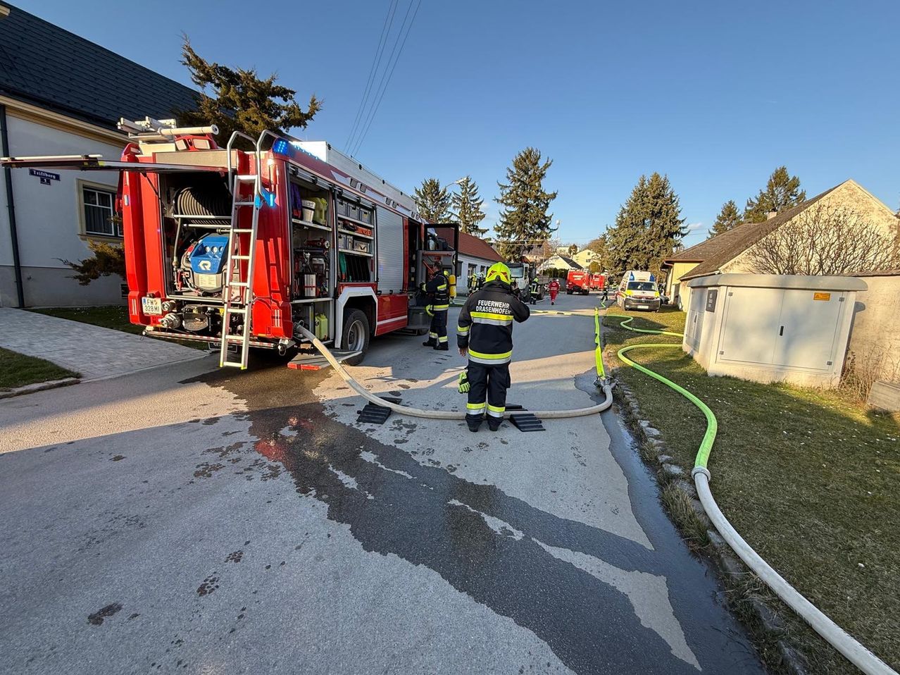 Ein Feuerwehrauto ist auf der Straße geparkt, vor dem ein Feuerwehrmann steht. Das Auto ist mit einem Schlauch verbunden, der Wasser sprüht.