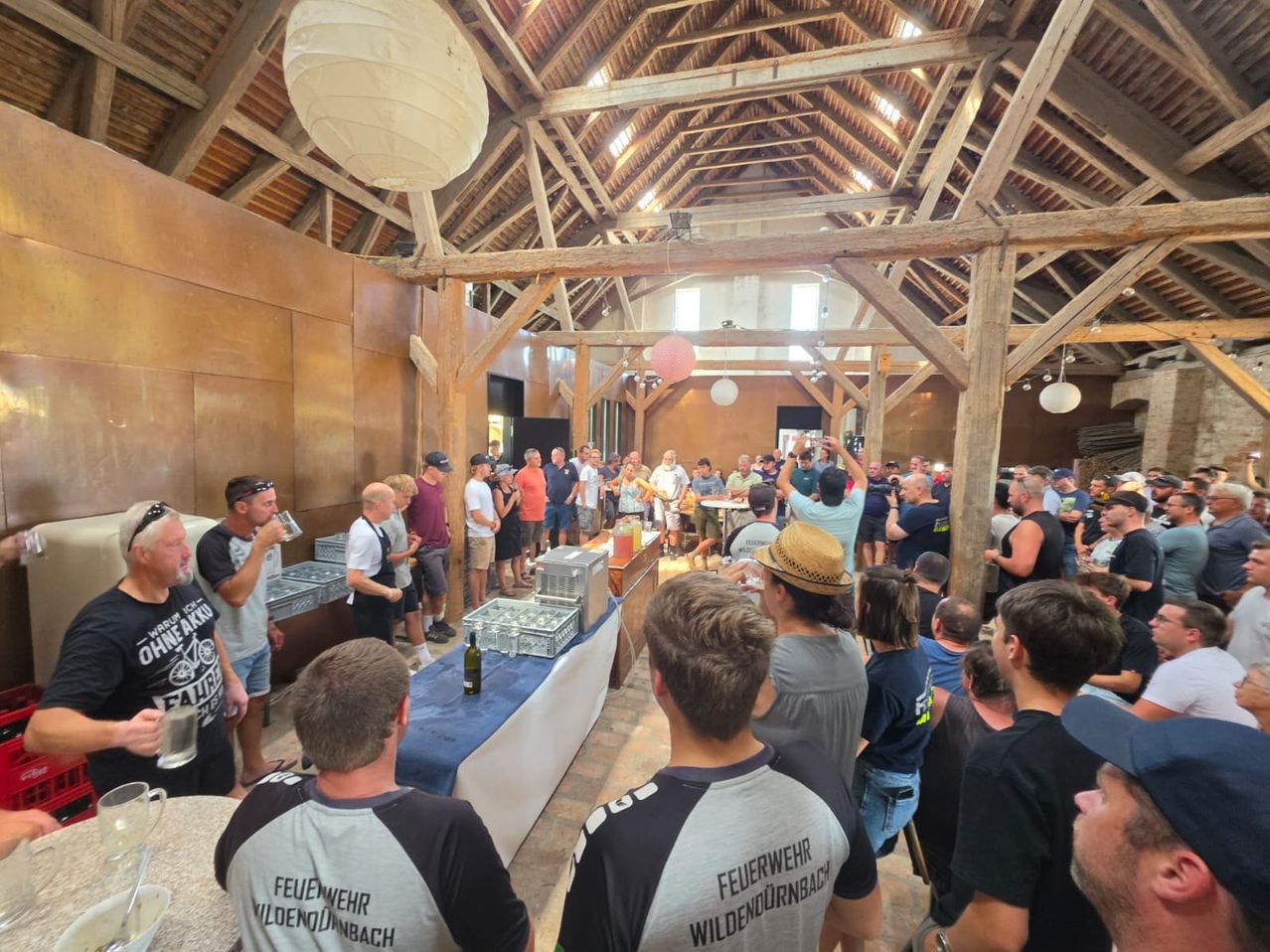 Eine große Gruppe von Menschen versammelt sich drinnen, wahrscheinlich bei einer sozialen Veranstaltung. Einige stehen, andere sitzen, und es gibt Tische mit verschiedenen Gegenständen darauf. Ein Mann in einem Hut hebt seine Hände.