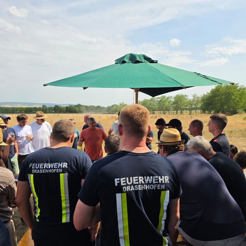 Eine Gruppe von Feuerwehrleuten in Uniform steht unter einem grünen Regenschirm auf einem offenen Feld. Einige tragen Hüte und andere halten Getränke. Im Hintergrund sind Bäume und eine entfernte Bergkette zu sehen.