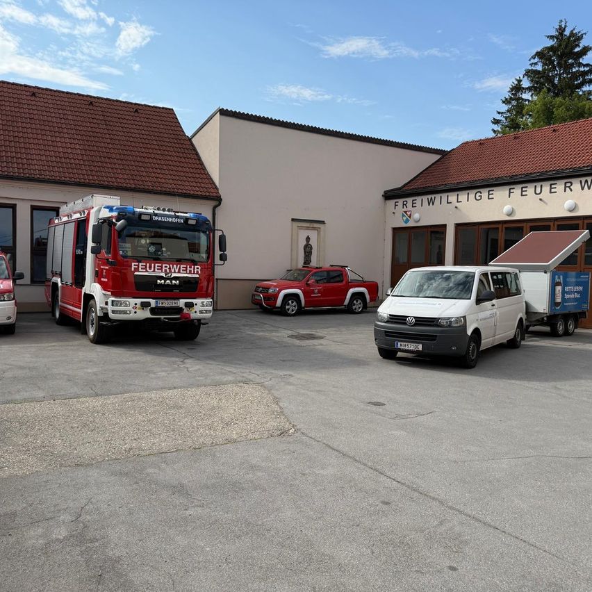 Ein Feuerwehrauto, ein roter Pickup-Truck und ein weißer Van sind vor einem Gebäude geparkt, auf dem der Text 'Freiwillige Feuerwehr' steht.