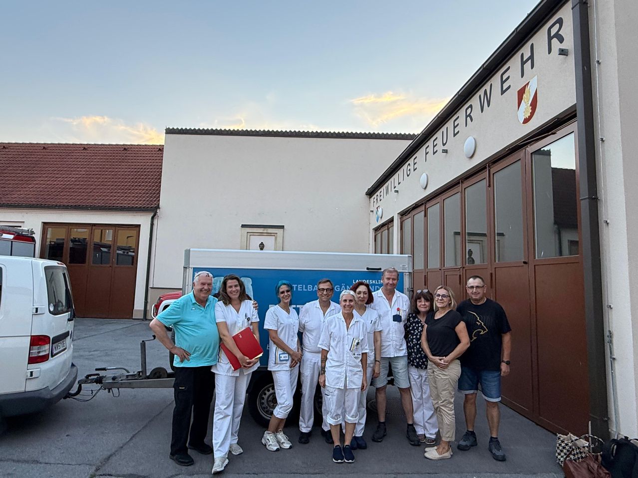 Eine Gruppe von Menschen, einige in weißen Uniformen, steht vor einem Gebäude mit einem Schild, das Freiwillige Feuerwehr liest. Ein blauer und weißer Anhänger ist hinter ihnen geparkt.