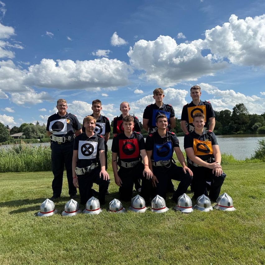 Eine Gruppe Feuerwehrleute in Uniform posiert für ein Foto vor einem See mit auf dem Rasen platzierten Helmen.