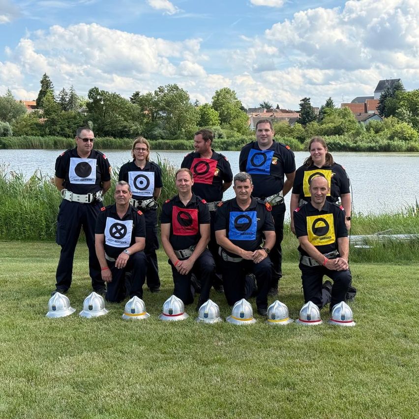 Eine Gruppe Feuerwehrleute in Uniform posiert mit Helmen auf dem Rasen in der Nähe eines Sees.