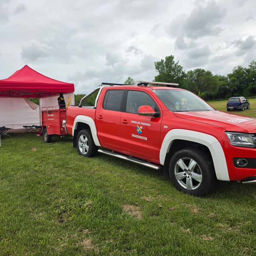 Ein roter Pickup-Truck mit weißen Akzenten steht auf einem Rasenfeld. Ein Zelt und ein roter Anhänger sind in der Nähe. Bäume und ein bewölkter Himmel sind im Hintergrund.