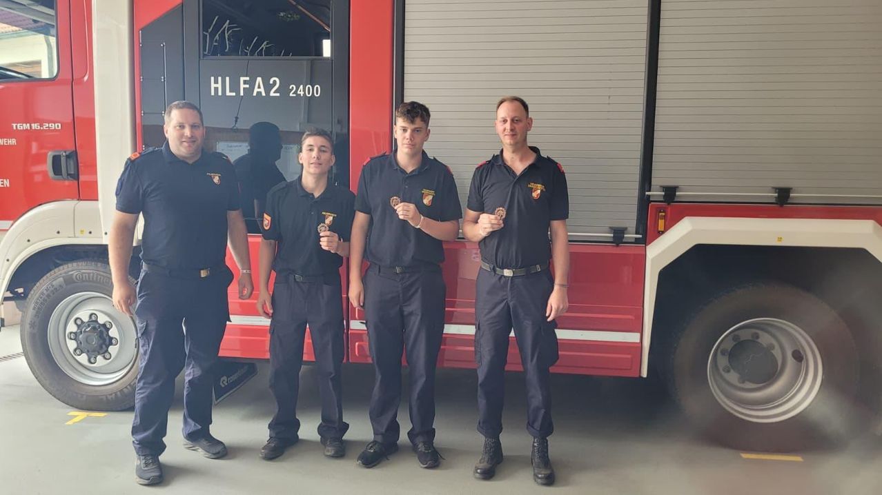 Four firefighters, wearing black uniforms and medals, stand in front of a red fire truck with 'HLF A2 2400' on the door.