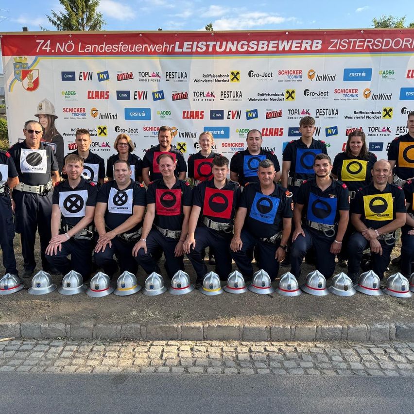 A group of firefighters, wearing helmets, uniforms, and glasses, poses for a photo in front of a large banner with logos and texts.