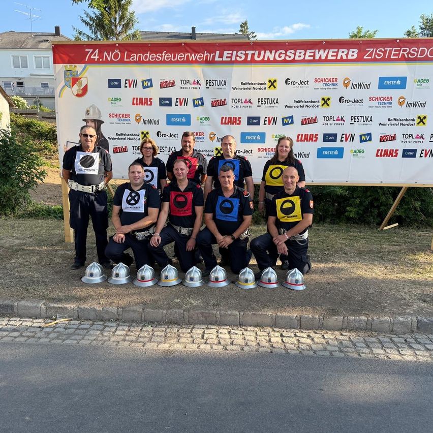 A group of firefighters pose for a photo in front of a large banner with various sponsors and logos. The firefighters are wearing uniforms and helmets.