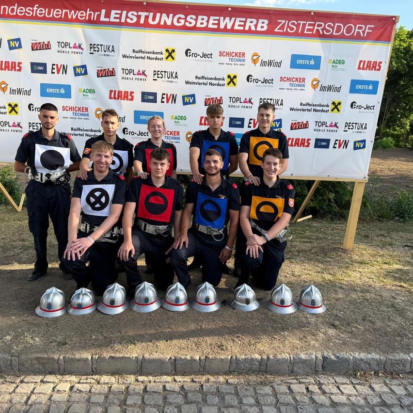 A group of firefighters in uniform poses for a photo in front of a banner with various logos, with helmets arranged in front of them.
