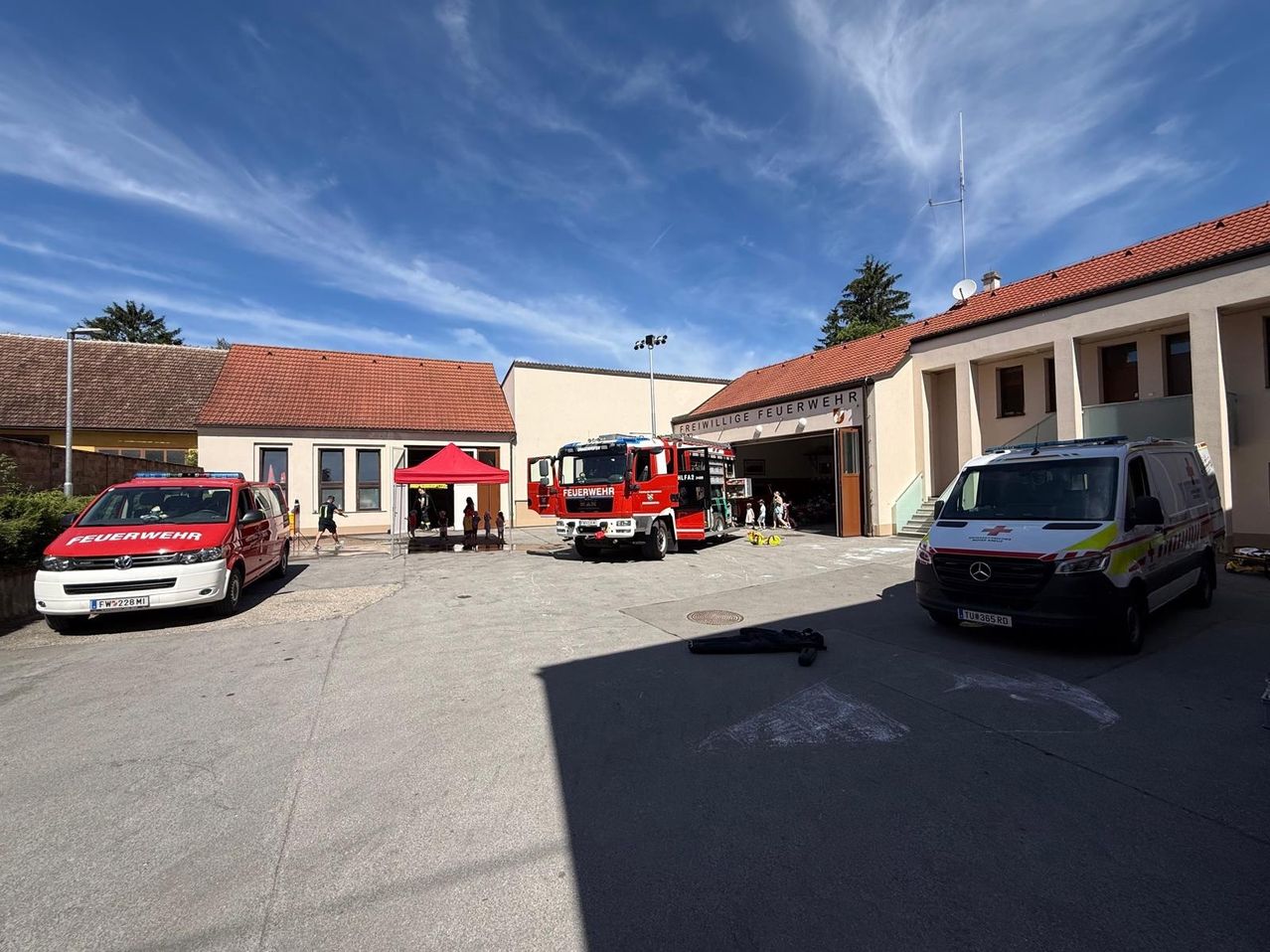 A fire station with a red fire truck and an ambulance parked outside. A tent is set up, and people are gathered around.