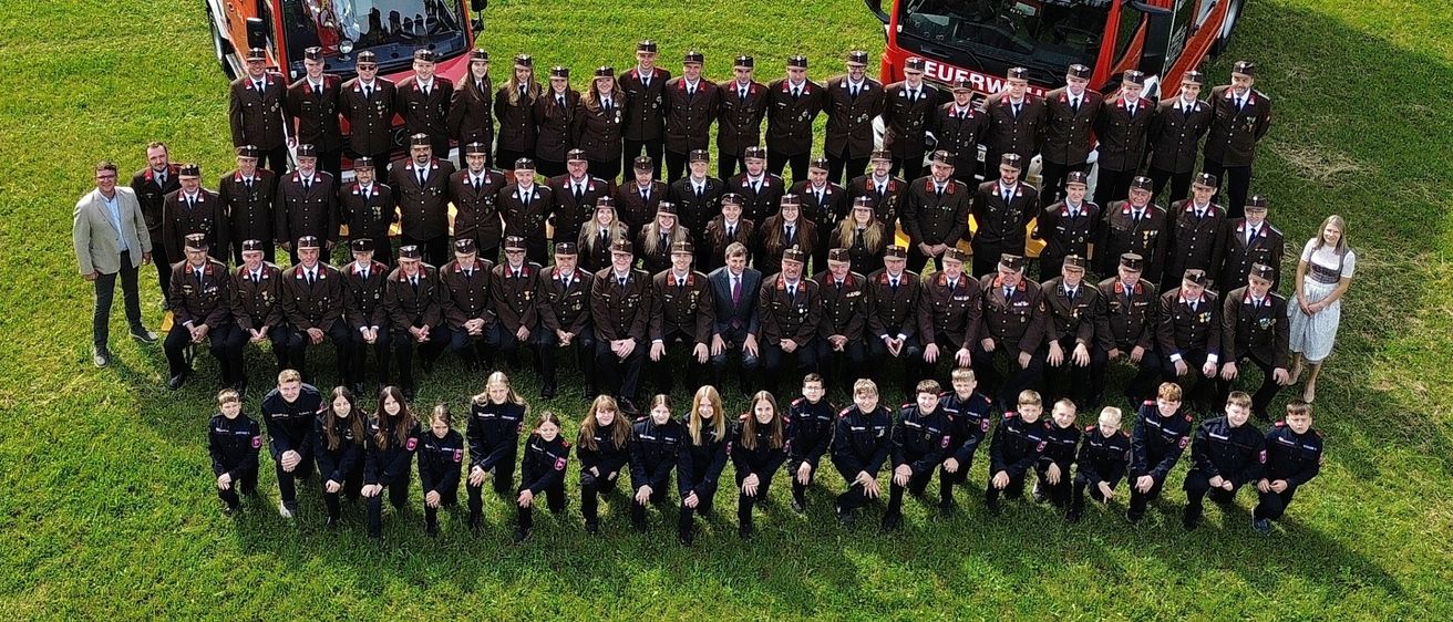 Eine Gruppe von Feuerwehrleuten posiert für ein Foto auf einem grünen Feld, vor zwei Feuerwehrautos, eines davon mit dem Wort Sand.