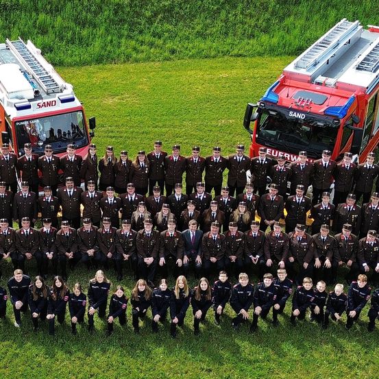 Eine große Gruppe von Feuerwehrleuten in Uniform posiert für ein Foto auf einem grünen Feld mit zwei Feuerwehrwagen im Hintergrund.