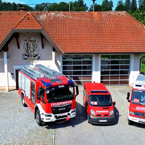 Drei Feuerwehrfahrzeuge sind vor einer Feuerwache mit Ziegeldach geparkt. Ein Fahrzeug hat das Wort "Feuerwehr" darauf.