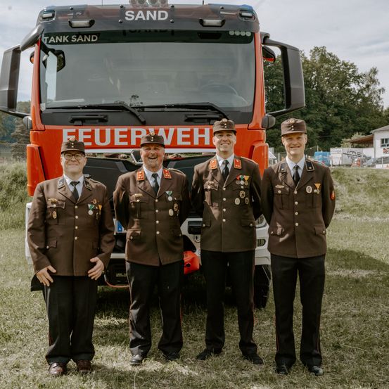 Vier Männer in Militäruniformen stehen vor einem roten Feuerwehrauto mit der Aufschrift 'Feuerwehr.' Sie lächeln und posieren wahrscheinlich für ein Foto. Im Hintergrund sind Bäume und ein Gebäude zu sehen.
