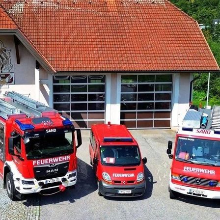 Drei rote Feuerwehrfahrzeuge sind vor einem Gebäude mit rotem Dach und großen Glastüren geparkt. Das Gebäude hat ein Logo an der Wand und ist von Grün umgeben.
