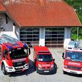 Drei rote Feuerwehrfahrzeuge sind vor einem Gebäude mit rotem Dach und großen Glastüren geparkt. Das Gebäude hat ein Logo an der Wand und ist von Grün umgeben.