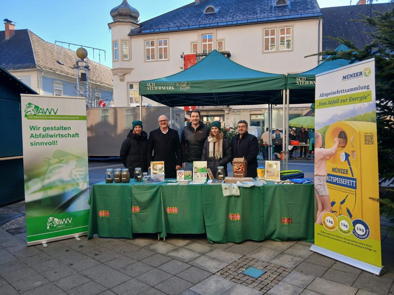 Fünf Personen stehen hinter einem mit grünem Tuch bedeckten Tisch, möglicherweise auf einem Markt. Dahinter ist ein großes grünes Zelt zu sehen.