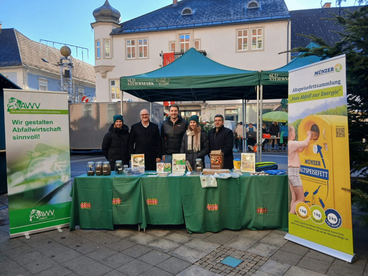 Fünf Personen stehen hinter einem mit einem grünen Tischtuch bedeckten Tisch und posieren für ein Foto. Hinter ihnen steht ein Zelt mit einer Bannerfahne. Sie stehen vor einem Gebäude mit vielen Fenstern.