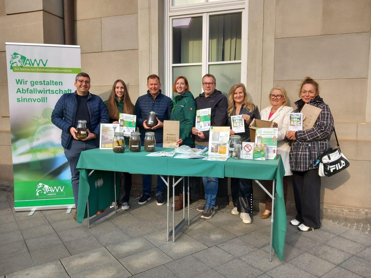 A group of people stand around a table with jars and pamphlets, possibly at an environmental event.
