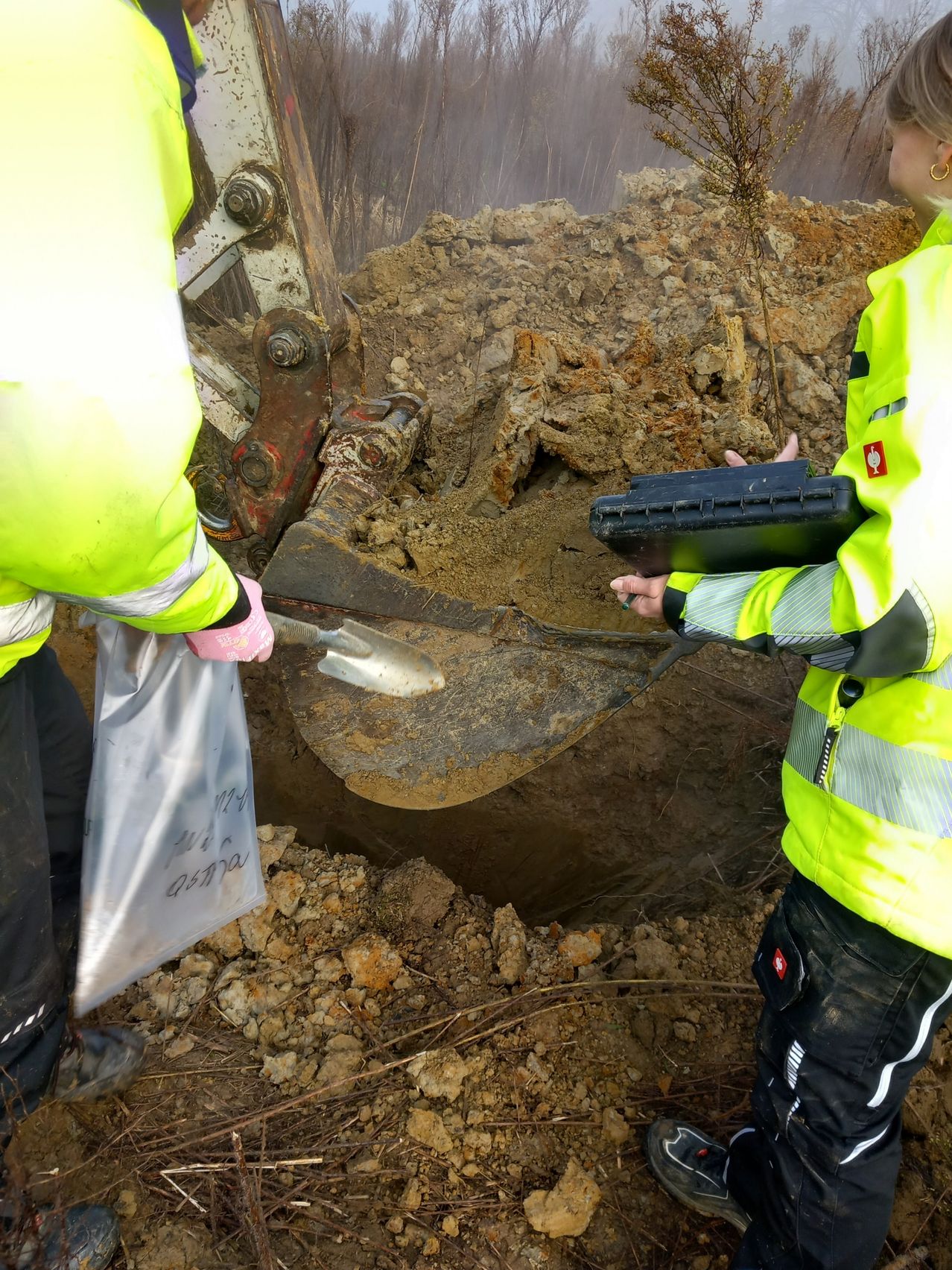 Two workers in neon vests are digging in dirt. One holds a plastic bag and the other a shovel. Nearby is a red logo.