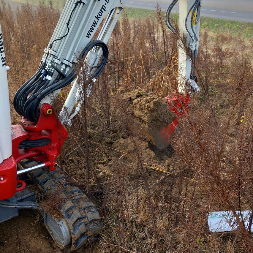 A red and white excavator is digging in the soil. It has many wires connected to it. A white pole is nearby.