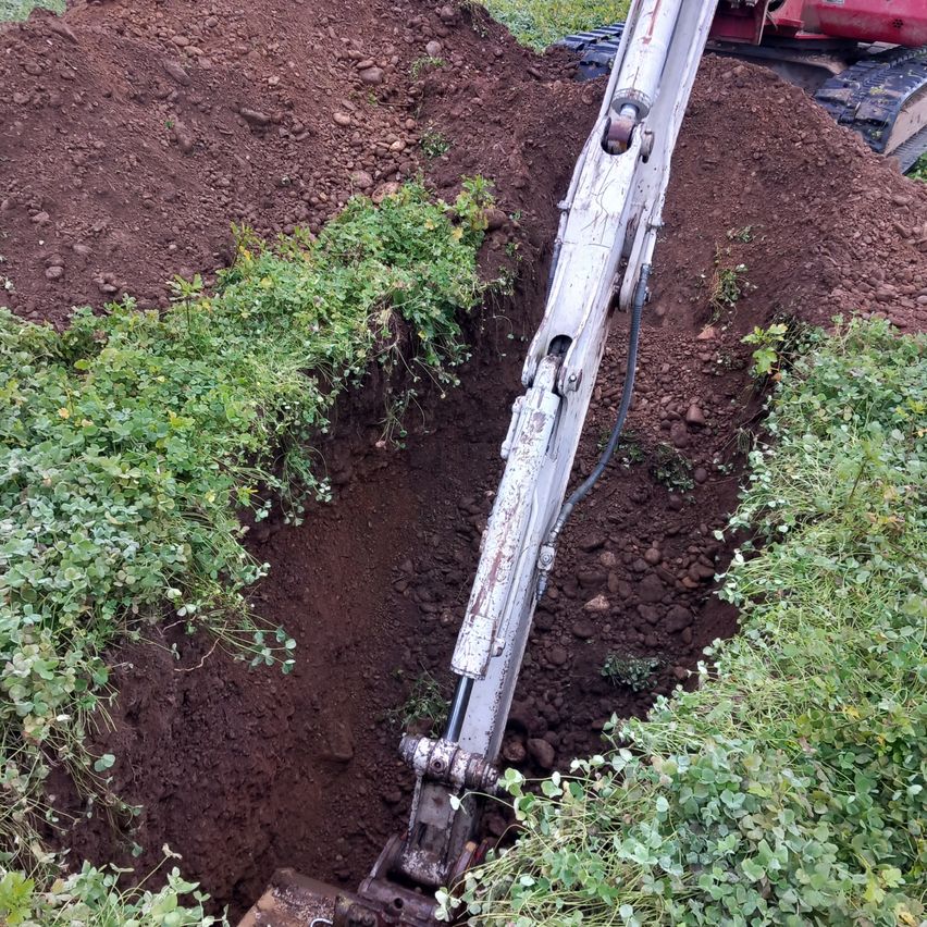A large digging machine is in the middle of a trench, with some plants growing on the sides. The soil is brown and rocky.