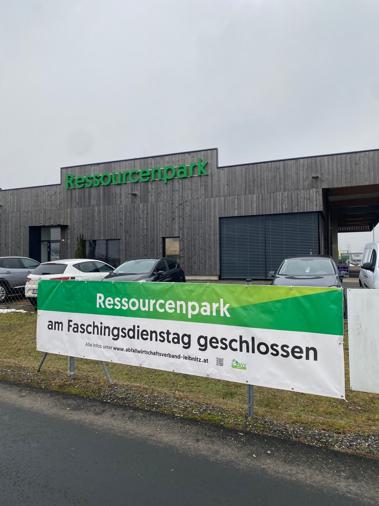 A closed signboard at Resourcepark is on the side of the road. Cars are parked in front of the building. The sky is cloudy.