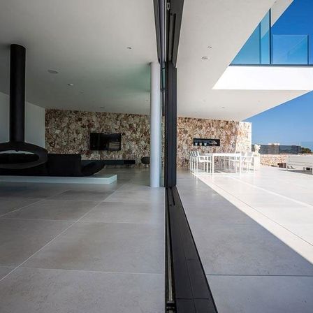 Ein modernes Haus mit großen Glasfenstern, einem Kamin und einem Essbereich mit weißen Stühlen und Tischen. Das Haus hat eine Steinwand und einen klaren blauen Himmel.