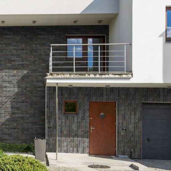 Ein modernes Haus mit einer Ziegelwand und einem Balkon. Es hat eine braune Tür und eine Garage. Der Balkon hat ein Metallgeländer.