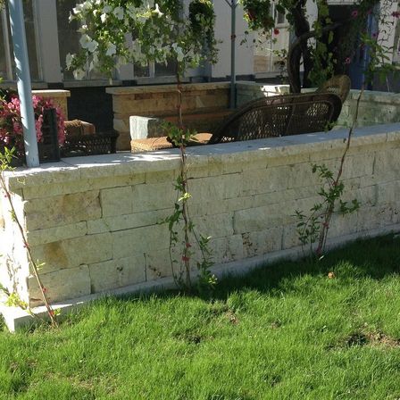 Ein Gartenbereich mit einer Steinmauer, grünen Pflanzen und einem Korbstuhl. Im Hintergrund befindet sich ein Gebäude mit Glasfenstern.