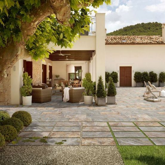 Eine weiße Villa mit einem Ziegeldach, umgeben von einem Innenhof mit Steinfliesen, Topfpflanzen und einem Baum. Es gibt Liegestühle, einen Tisch und eine Couch auf dem Innenhof.