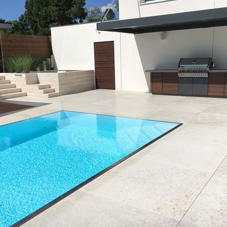 Ein Außenpool ist in ein modernes Haus integriert. Der Pool ist von einer weißen Betonterrasse umgeben und hat einen schwarzen Rand. Rechts befindet sich ein eingebauter Grill unter einem Vordach.