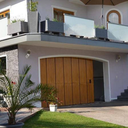 Ein modernes Haus mit Balkon und einer hölzernen Garagentür. Der Balkon hat Topfpflanzen und einen geschlossenen Schirm. Das Haus hat ein Glasfenster und eine Steinmauer.