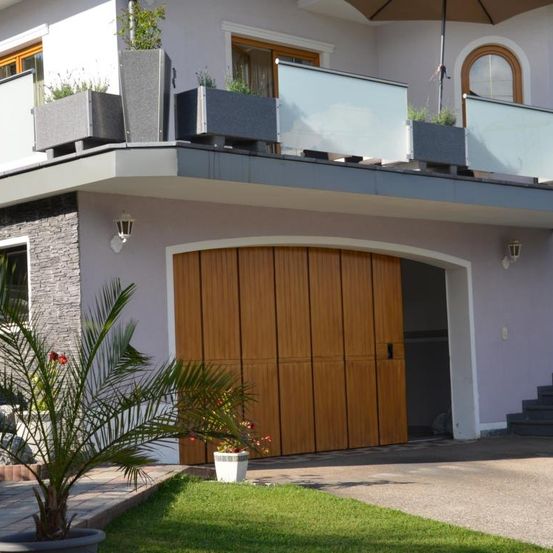 Ein modernes Haus mit Balkon und einer hölzernen Garagentür. Der Balkon hat Topfpflanzen und einen geschlossenen Schirm. Das Haus hat ein Glasfenster und eine Steinmauer.