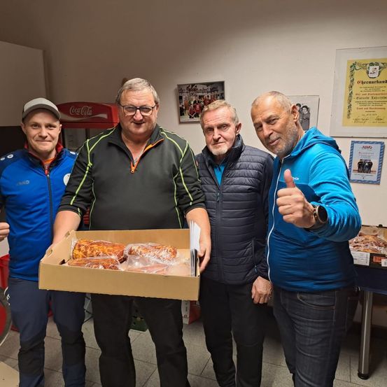 Vier Männer stehen in einem Raum mit einem Coca-Cola-Automaten im Hintergrund. Sie halten eine Schachtel mit Essen. Ein Mann gibt einen Daumen hoch.