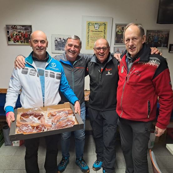 Vier Männer stehen in einem Raum mit weißer Wand zusammen. Ein Mann hält eine Schachtel mit eingewickeltem Essen. Der Mann in der Mitte trägt eine Brille und eine graue Jacke. Die anderen Männer tragen Jacken mit einem Logo auf der linken Seite.