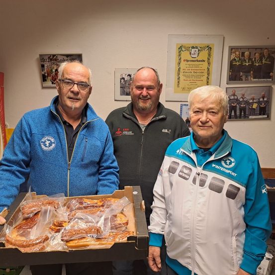 Drei Männer stehen in einem Raum mit einer weißen Wand. Sie lächeln und scheinen für ein Foto zu posieren. Der Mann auf der linken Seite hält eine Schachtel mit einem Sandwich und der Mann in der Mitte hält ein Stück Papier.