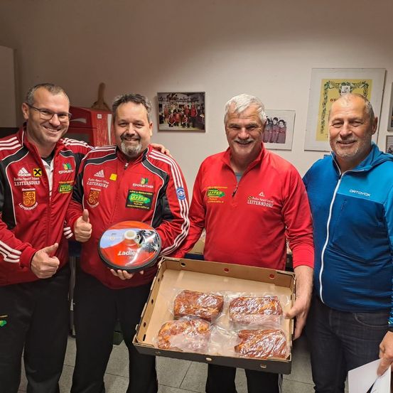 Vier Männer stehen nebeneinander in einem Raum. Der Mann in der Mitte hält eine Schachtel mit Essen.