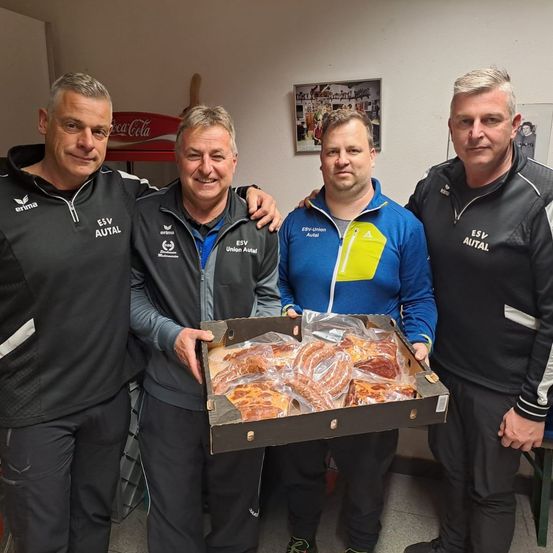 Vier Männer in Sportbekleidung stehen in einem Raum mit einem Coca-Cola-Schild an der Wand. Ein Mann hält eine Schachtel mit eingewickeltem Essen.