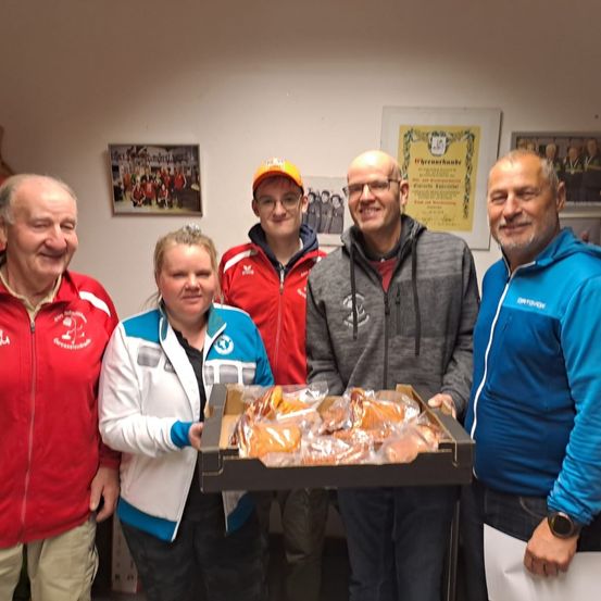 Fünf Personen stehen zusammen in einem Raum. Sie lächeln und posieren für ein Foto. Eine Person hält eine Box mit Essen. An der Wand hinter ihnen hängen Bilderrahmen.