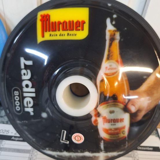 Ein schwarzes Plastiketikett mit dem Markennamen Murauer ist abgebildet. Es hat einen gelben Aufkleber mit dem Text 'Rein das Beste'. Das Etikett zeigt auch eine Flasche Murauer Bier mit einer Hand, die es hält.